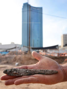 Bronze dagger found, against the backdrop of the excavation and the towers. (Yoli Shwartz/IAA)